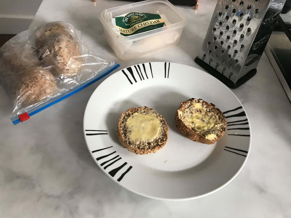One toasted roll split in half and buttered on white plate. Next to the plate is a cheese grater, a tupperware containing a packet of mature Lye Cross Farm cheddar cheese and a ziplock bag containing a couple of essential bread rolls just out of the freezer. 