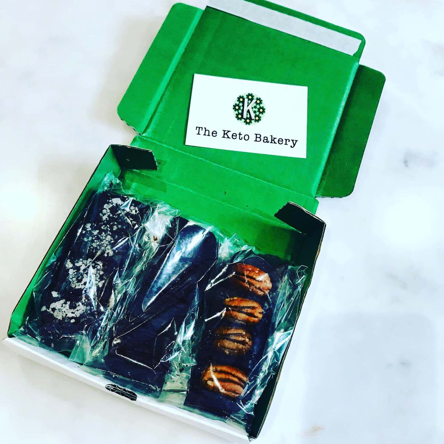 green card posting box of 12 Chocketos - brownie bites from The Keto Bakery wrapped in cellophane.. 4 are topped with pecan slices, 4 are topped with seasalt sprinkle. There is a small white card with The Keto bakery logo inside the box.