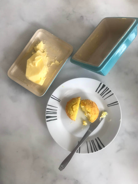 Turmeric muffin from the Keto Bakery split in half on white plate with knife and butter. Next to the plate is an aqua blue coloured open butter dish containing a large slab of butter.